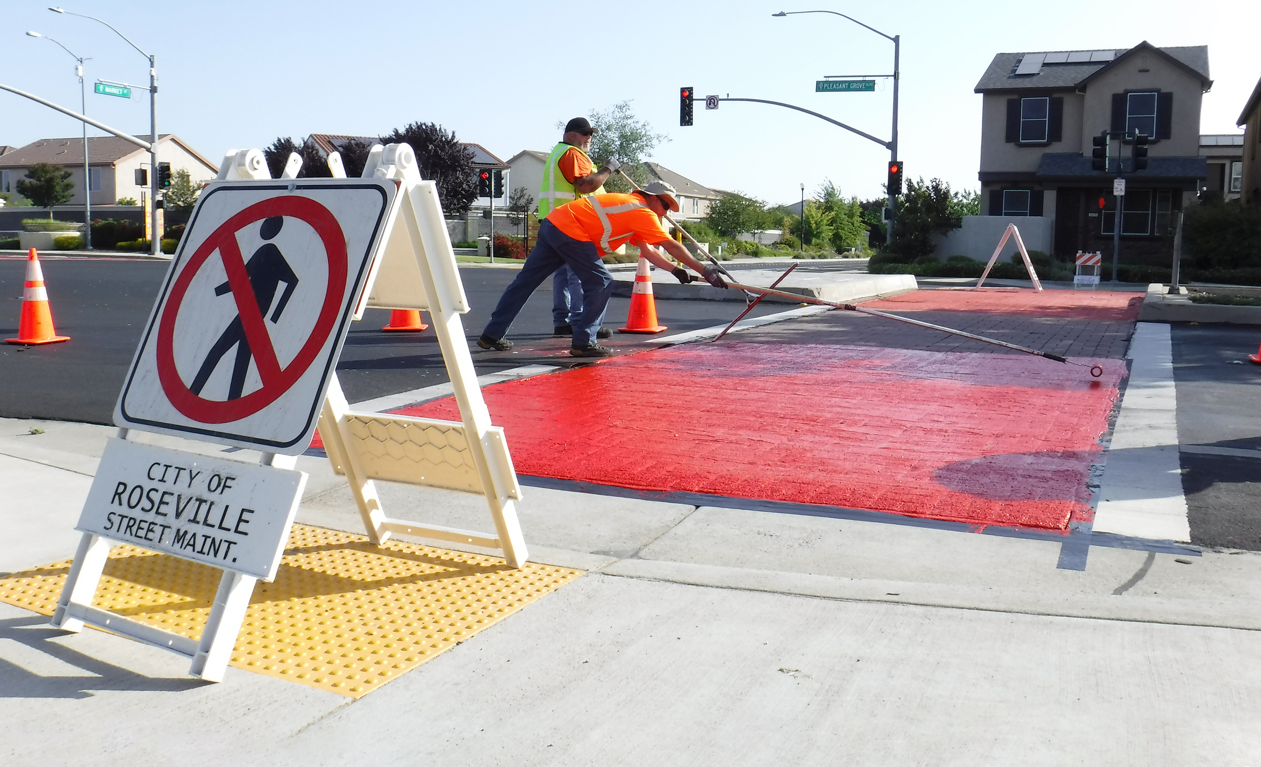 Workers apply red paint to a crosswalk, with traffic cones and a 